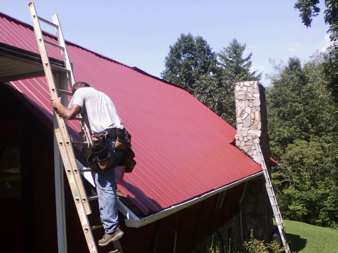 Roofer on a ladder beside a red metal roof and stone chimney — Soffit & Fascia Repair by Roofing Brothers in Curtis Bay
